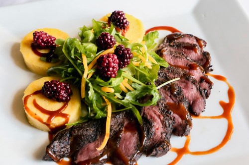 A plate featuring sliced steak, polenta rounds, arugula salad, and blackberries drizzled with sauce.