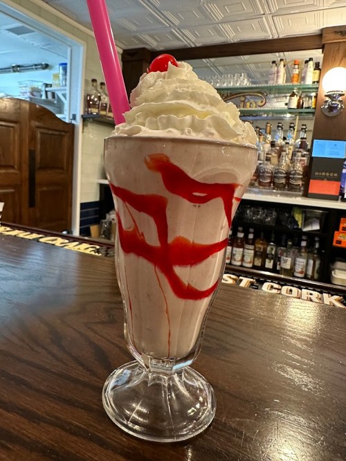 A tall glass of milkshake topped with whipped cream, a cherry, and drizzled with red syrup, sitting on a wooden counter.