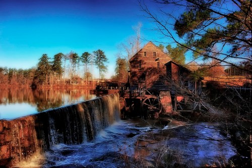 A historic watermill beside a calm lake, with a waterfall cascading over rocks and surrounded by trees.