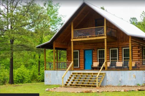 A rustic wooden cabin with a blue door, surrounded by greenery and featuring a porch with steps.