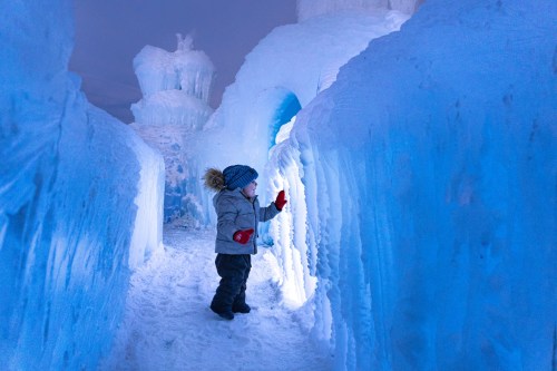 A child in winter clothing touches a glowing ice wall in a snowy, icy landscape.
