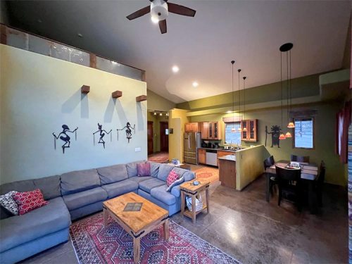 Cozy living room with a sectional sofa, wooden table, and kitchen area, featuring decorative wall art and warm lighting.
