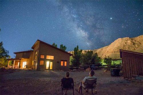 Two people sit outside a house, gazing at a starry night sky filled with the Milky Way and distant mountains.