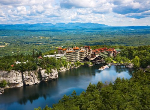 A scenic view of a lakeside resort surrounded by lush greenery and mountains under a partly cloudy sky.
