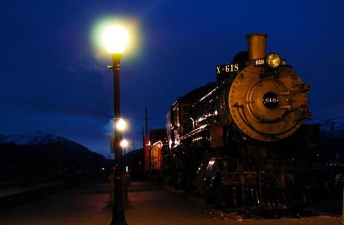 A vintage steam train illuminated by street lamps at dusk, with mountains in the background.