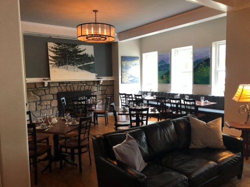 Cozy restaurant interior with wooden tables, a stone fireplace, and large windows, featuring artwork on the walls.