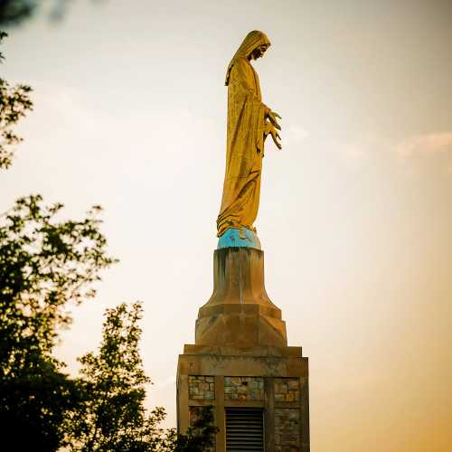 A statue of a figure with outstretched arms, set against a sunset sky, surrounded by trees.