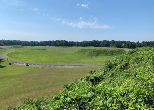 A wide view of a grassy landscape with a road, trees in the background, and a large earthen mound.