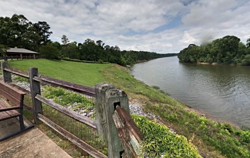 A serene riverbank scene with lush greenery, a wooden fence, and a cloudy sky reflecting over the calm water.
