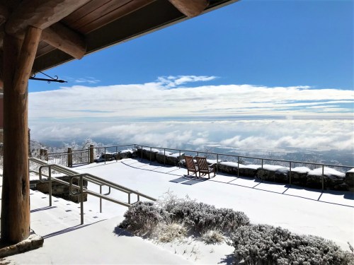 Snow-covered patio with wooden benches, overlooking a cloudy valley under a bright blue sky.