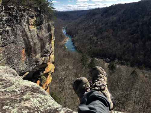A person’s feet in hiking boots resting on a rocky ledge, overlooking a river and forested valley below.