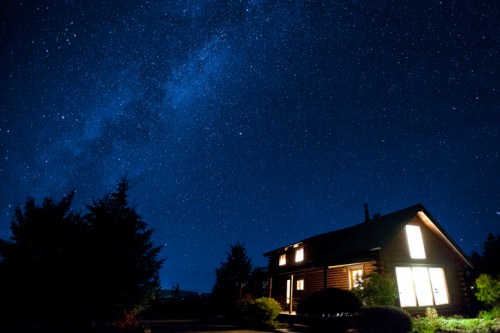 A cozy log cabin illuminated at night, surrounded by trees under a starry sky with the Milky Way visible.