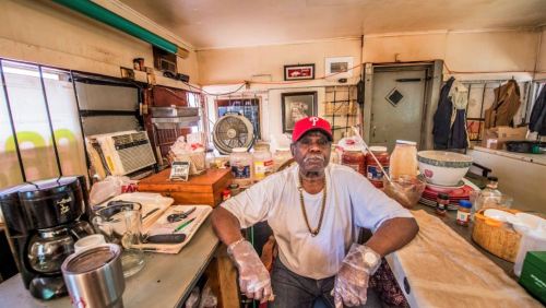 A man in a red cap sits in a kitchen filled with cooking supplies and ingredients, wearing gloves and a relaxed expression.