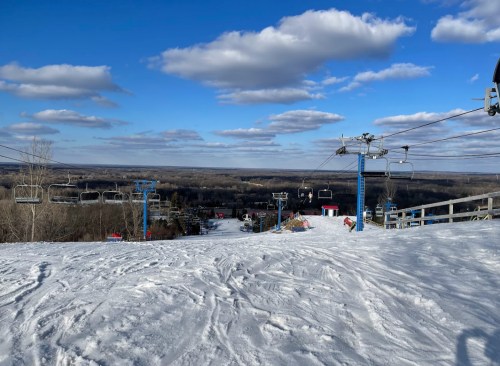 A snowy ski slope with chairlifts and a scenic view of the landscape under a blue sky with fluffy clouds.