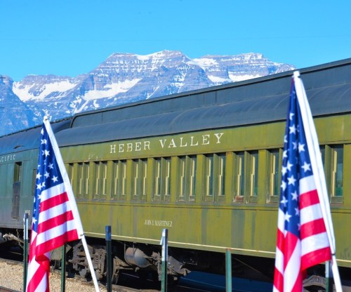 A vintage train car labeled "Heber Valley" with American flags in the foreground and mountains in the background.