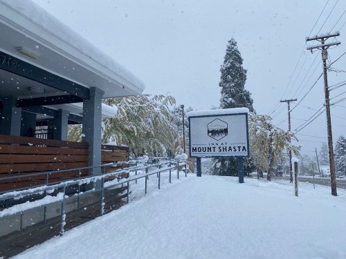 A snowy scene featuring the sign for the Inn at Mount Shasta, with snow-covered ground and trees in the background.