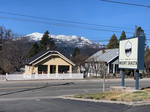 A sign for "Live at Mount Shasta" in front of a house with snow-capped mountains in the background.