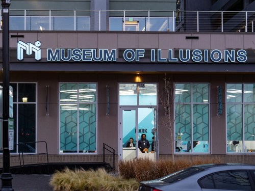 Exterior view of the Museum of Illusions, featuring a modern sign and large windows with people inside.