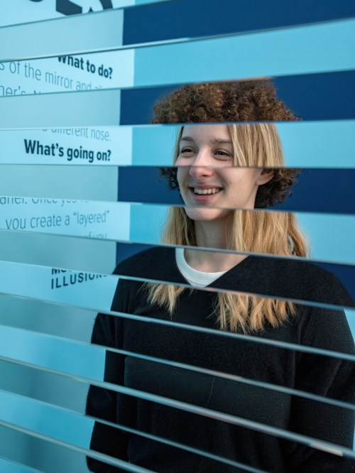 A smiling woman with long hair stands behind horizontal slats, with a blue wall and text in the background.