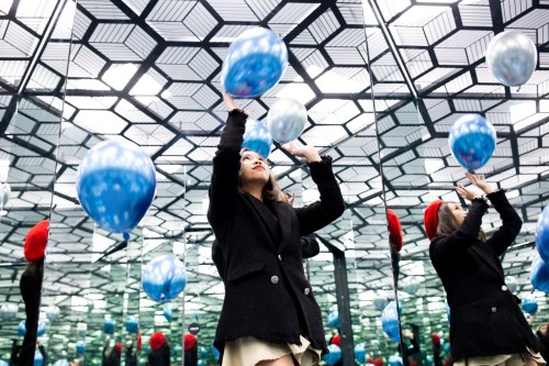 A woman in a black coat reaches for blue balloons in a mirrored room with geometric patterns.