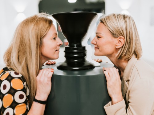 Two women lean in closely, smiling at each other while facing a black sculpture between them.