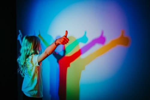 A child with long hair gives a thumbs-up, casting colorful shadows on the wall.