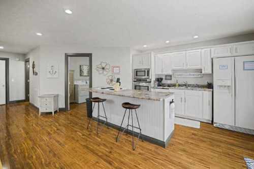 Modern kitchen with white cabinets, granite island, and wooden floors, featuring bar stools and stainless steel appliances.