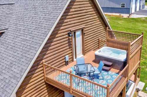 Aerial view of a wooden cabin with a deck featuring a hot tub and outdoor seating on a sunny day.