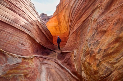 A person stands in a narrow, colorful sandstone canyon with swirling patterns and warm hues of orange and red.