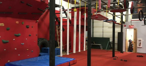 Indoor gym with a red climbing wall, hanging obstacles, and a blue mat on the floor.