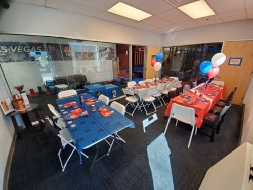 A decorated party room with tables set in red and blue, balloons, and a view of a gym area through glass walls.