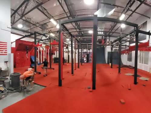 A spacious indoor gym with red flooring and various climbing and training equipment. People are engaged in workouts.