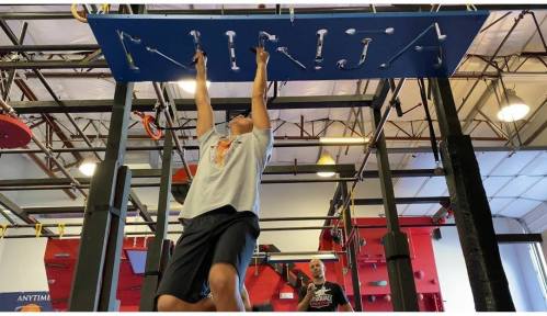 A person hangs from a blue obstacle labeled "NINJA" in a gym, with another individual in the background.