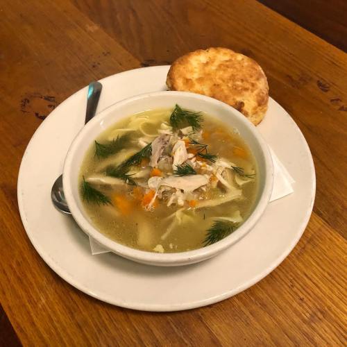 A bowl of chicken soup with vegetables and herbs, served with a biscuit on the side, on a wooden table.