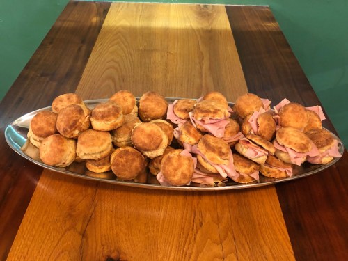 A silver platter filled with small biscuits and ham sandwiches, arranged neatly on a wooden table.