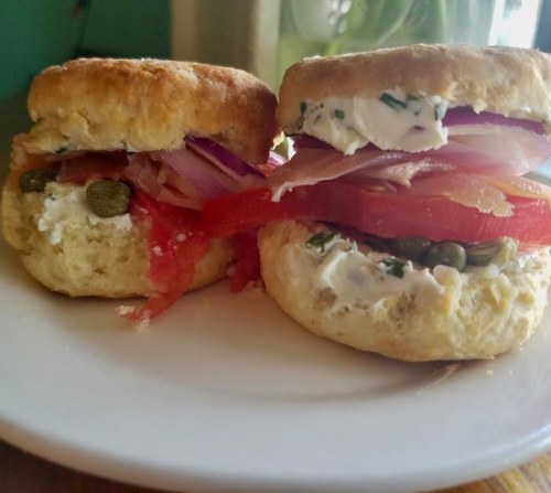 Two biscuit sandwiches filled with cream cheese, bacon, tomato, red onion, and capers, served on a white plate.