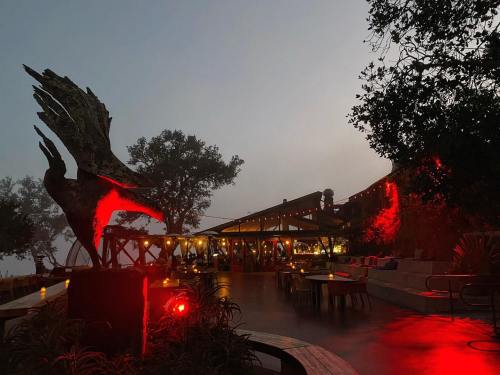 A misty evening scene at a restaurant with a large bird sculpture and red lighting, surrounded by trees.