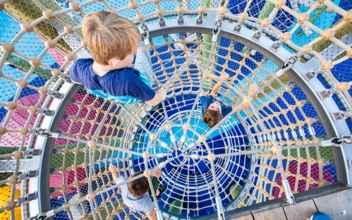 Children climbing a spiral net structure in a colorful playground, viewed from above.