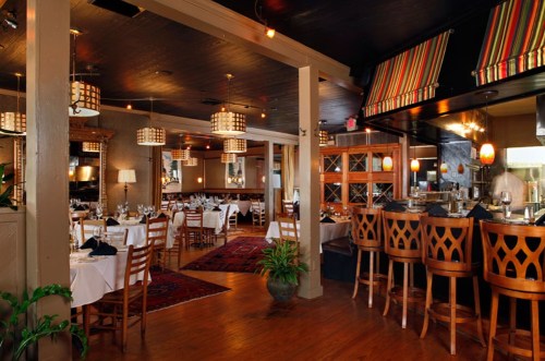 Cozy restaurant interior with wooden tables, chairs, and a bar, featuring warm lighting and decorative accents.