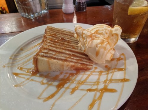 A slice of caramel drizzled cake with a scoop of vanilla ice cream on a white plate, next to a drink with a lemon slice.