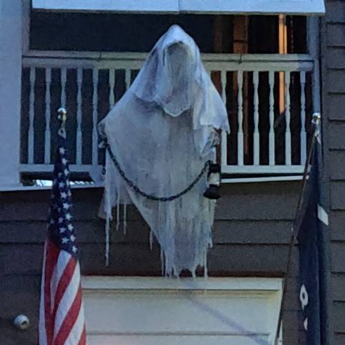 A ghostly figure draped in white hangs from a balcony, with American flags on either side.