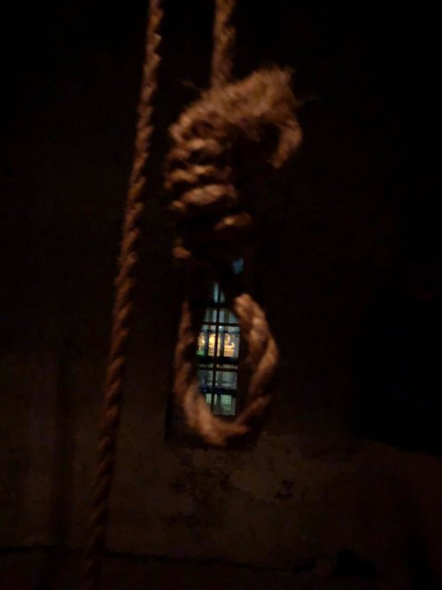 A dimly lit room featuring a noose hanging from a rope, with a faintly illuminated window in the background.