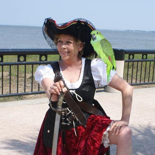 A woman in a pirate costume poses with a sword and a green parrot on her shoulder by the water.