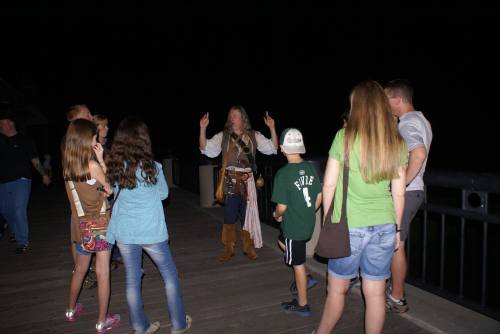 A storyteller in pirate attire engages a group of children and adults on a darkened pier.