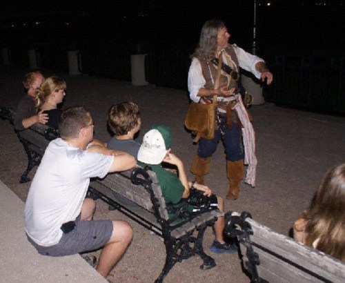 A storyteller in period costume engages a small audience seated on a bench at night.