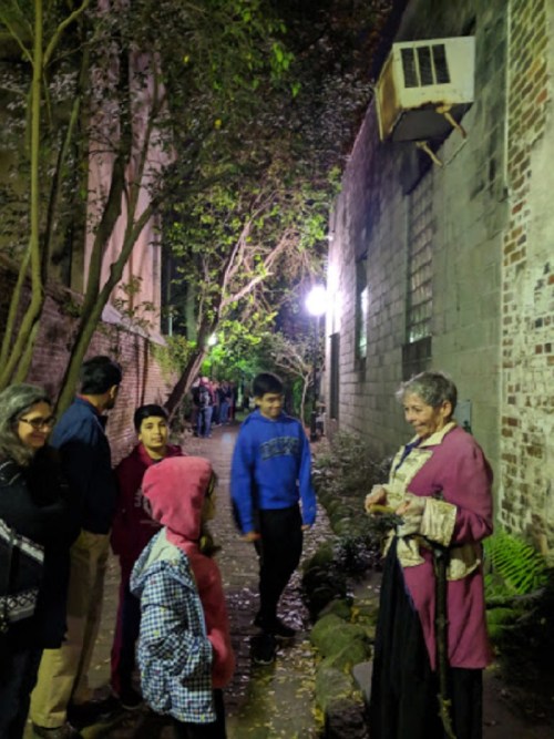 A group of people, including children, gather in a dimly lit alley, chatting with a woman in historical attire.
