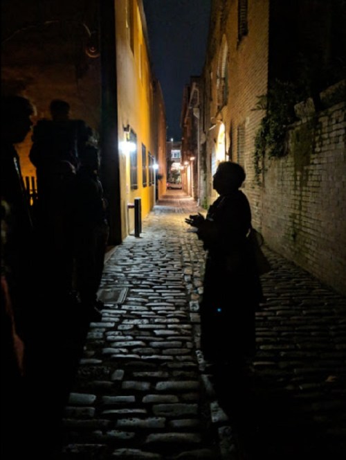 A silhouette of a person speaking in a dimly lit cobblestone alley, with shadows of others nearby.