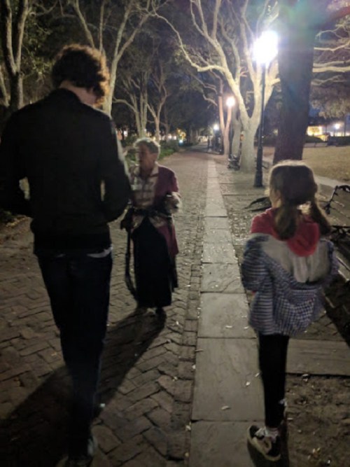 A man and a girl walk past an elderly woman in a park at night, illuminated by streetlights.