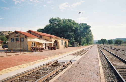 A small train station with a red-tiled roof, surrounded by trees and empty railway tracks under a clear blue sky.