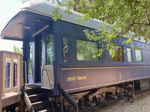 A vintage train car with a blue exterior, featuring large windows and a staircase, surrounded by greenery.